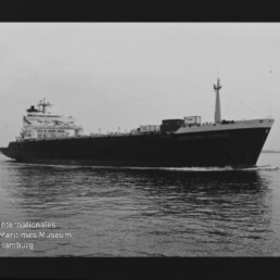 Schwarzweiß-Foto eines großen Frachtschiffs in Seitenansicht, mit Brückenaufbau achtern, Containern auf dem Deck und leichtem Heckwellengang auf ruhigem Wasser; schwarzer Filmrand mit Ziffer 15 und Museumslogo unten links.