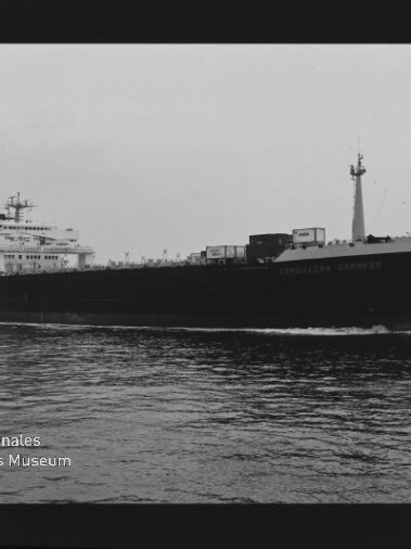 Schwarzweiß-Foto eines großen Frachtschiffs in Seitenansicht, mit Brückenaufbau achtern, Containern auf dem Deck und leichtem Heckwellengang auf ruhigem Wasser; schwarzer Filmrand mit Ziffer 15 und Museumslogo unten links.