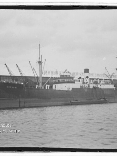 Schwarz‑weiß-Foto eines großen Frachtschiffs mit Masten und Schornstein, vertäut am Kai vor einem Lagerhaus mit mehreren Ladekränen, Wasser im Vordergrund und einem 'Internationales Maritimes Museum Hamburg'-Wasserzeichen unten links.
