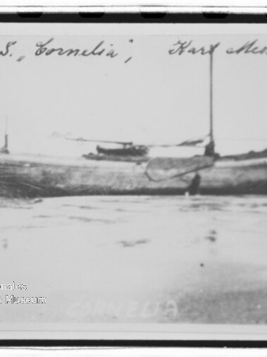 Schwarzweiß-Fotografie eines hölzernen Segelboots namens „Cornelia“, seitlich in seichtem Wasser oder am Strand liegend, mit aufragendem Mast, handschriftlicher Beschriftung am oberen Rand und Museumswasserzeichen unten links.