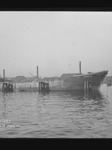 Schwarzweiß-Foto eines länglichen Frachtschiffs, das ruhig zwischen hölzernen Dalben in einem Industriehafen liegt, vor niedrigen Lagergebäuden im Hintergrund und mit dem Schriftzug des Internationalen Maritimen Museums Hamburg in der unteren linken Ecke.