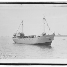Schwarzweiß-Foto eines weißen, zweimastigen Schiffs mit geschmückten Wimpelketten, das auf ruhigem Wasser vor einer flachen Küstenlinie liegt; links unten ein Wasserzeichen des Internationalen Maritimen Museums Hamburg.