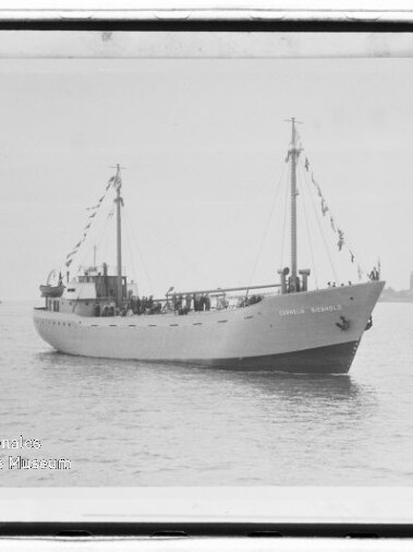 Schwarzweiß-Foto eines weißen, zweimastigen Schiffs mit geschmückten Wimpelketten, das auf ruhigem Wasser vor einer flachen Küstenlinie liegt; links unten ein Wasserzeichen des Internationalen Maritimen Museums Hamburg.