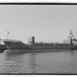 Schwarzweißaufnahme eines seitlich gezeigten Frachtschiffs namens 'CORINNA' im Hafen vor Ladekränen und einem Flutlichtmast, mit ruhiger Wasserfläche und dem Logo des Internationalen Maritimen Museums Hamburg links unten.