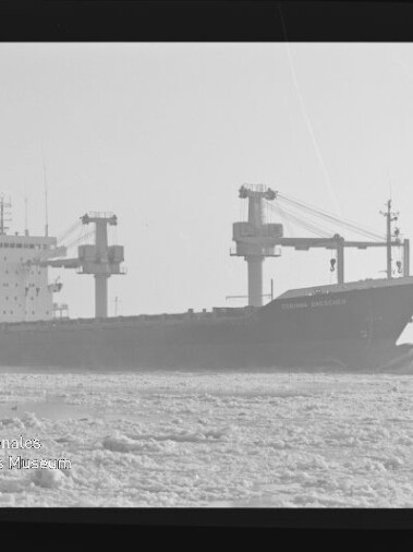 Schwarzweiß-Fotografie eines Frachtschiffs mit mehreren Deckskranen, das als dunkle Silhouette durch eisbedecktes Wasser fährt; am Bildrand sind Filmrandbeschriftungen (AGFAPAN 100) sichtbar.