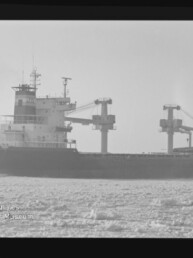 Schwarzweiß-Foto eines Frachtschiffs mit hoher Aufbautenbug und zwei großen Ladekränen, das in einer von Eisfeldern bedeckten See fährt; unten links sichtbar das Logo und der Schriftzug des Internationalen Maritimen Museums Hamburg.