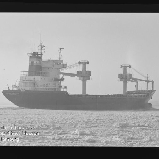 Schwarzweiß-Foto eines Frachtschiffs mit hoher Aufbautenbug und zwei großen Ladekränen, das in einer von Eisfeldern bedeckten See fährt; unten links sichtbar das Logo und der Schriftzug des Internationalen Maritimen Museums Hamburg.