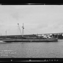 Schwarzweißaufnahme eines mittleren Frachtschiffs in Profilansicht mit zwei Masten und Aufbau, das auf ruhigem Wasser fährt; im Hintergrund Ufer mit Bäumen und einer langen Eisenbahnbrücke, unten links Museumswasserzeichen.