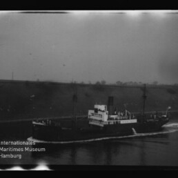 Schwarzweiß-Foto eines dampfbetriebenen Frachtschiffs mit Schornstein, zwei Masten und deutlich sichtbarer Heck- und Bugwelle, das in einem Kanal oder Fluss entlang einer niedrigen, baumbestandenen Uferböschung fährt; unten links Museumslogo und Schriftzug Internationales Maritimes Museum Hamburg.
