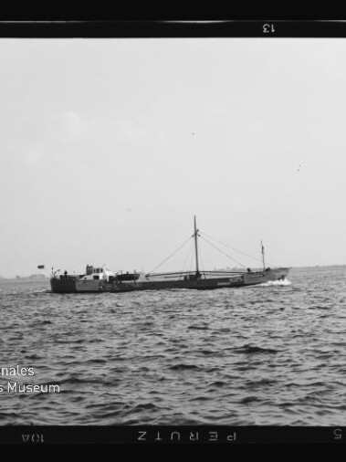 Schwarzweißaufnahme eines flachen Frachtschiffs mit mittigem Mast und Ladebaum, das auf ruhiger See nahe dem Horizont fährt; im Vordergrund wellige Wasseroberfläche und unten links ein Wasserzeichen des Internationalen Maritimen Museums Hamburg.