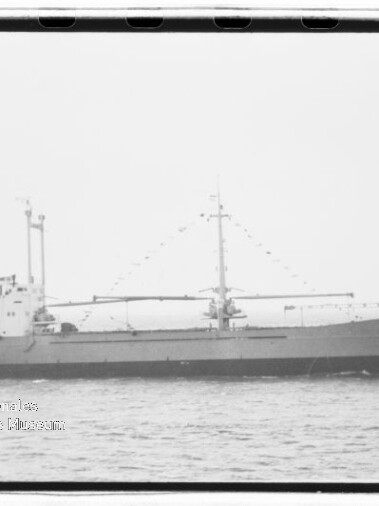 Schwarz-weiß-Seitenansicht eines Frachtschiffs mit flachem Deck, zentralem Mast und zwei Ladebäumen, das ruhig auf dem Meer fährt.