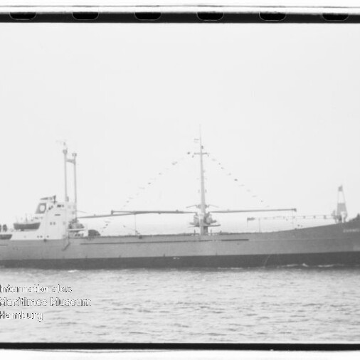 Schwarz-weiß-Seitenansicht eines Frachtschiffs mit flachem Deck, zentralem Mast und zwei Ladebäumen, das ruhig auf dem Meer fährt.