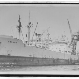 Schwarzweißfoto des Frachtschiffs CORONA, längsseits an einem Industriepier mit mehreren Ladekränen und Verladearmen, mit einem Schlepper am Heck und dem Logo des Internationalen Maritimen Museums Hamburg unten links.