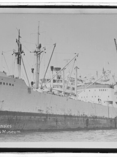 Schwarzweißfoto des Frachtschiffs CORONA, längsseits an einem Industriepier mit mehreren Ladekränen und Verladearmen, mit einem Schlepper am Heck und dem Logo des Internationalen Maritimen Museums Hamburg unten links.