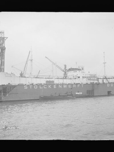 Großes Frachtschiff in einem Dock, an der Dockwand die Aufschrift STÜLCKENWERFT DOCK VI, mit mehreren Kränen und Hafenanlagen im Hintergrund und Wasser im Vordergrund.