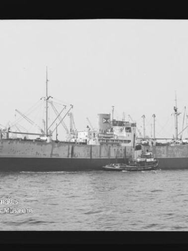 Schwarz‑weiß-Foto eines großen Frachtschiffs mit mehreren Masten und Ladekränen, begleitet von einem kleinen Schlepper auf dem Wasser; Filmrand und ein Wasserzeichen des Internationalen Maritimen Museums Hamburg links unten sind sichtbar.
