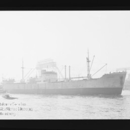 Schwarz-weiß-Aufnahme eines großen Frachtschiffs in Seitenansicht im Hafen, mit Ladekränen und Masten, Industrieanlagen im Hintergrund, einem kleinen Beiboot am Bug und sichtbarem schwarzen Filmrand