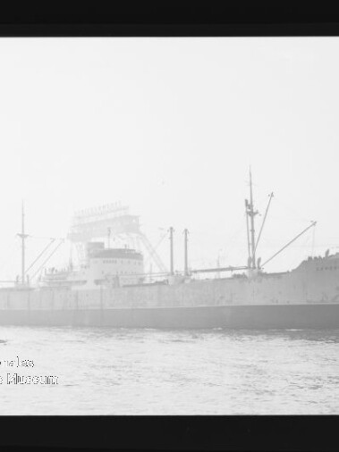Schwarz-weiß-Aufnahme eines großen Frachtschiffs in Seitenansicht im Hafen, mit Ladekränen und Masten, Industrieanlagen im Hintergrund, einem kleinen Beiboot am Bug und sichtbarem schwarzen Filmrand