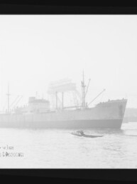 Großes Frachtschiff in einem nebligen Hafen, im Vordergrund ein kleines Motorboot und im Hintergrund Kräne und Werftanlagen, schwarz-weiß aufgenommen.