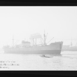 Großes Frachtschiff in einem nebligen Hafen, im Vordergrund ein kleines Motorboot und im Hintergrund Kräne und Werftanlagen, schwarz-weiß aufgenommen.