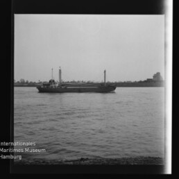Schwarz‑weiß-Aufnahme eines Frachtschiffs mit zwei Masten, das ruhig auf einem breiten Fluss vor einer niedrig bebauten Uferlinie fährt; das Bild zeigt dunkle Negativränder und das Wasserzeichen des Internationalen Maritimen Museums Hamburg.