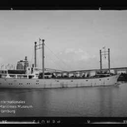 Schwarzweißaufnahme eines mittleren Frachtschiffs mit zwei Masten und Ladebäumen, das ruhig auf einem Fluss unter einer stählernen Brücke vorbeifährt, am Heck weht eine Flagge.