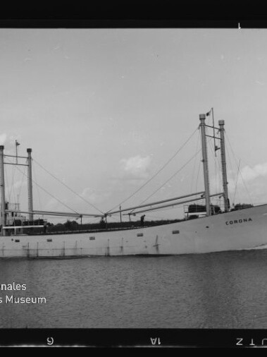Schwarzweißaufnahme eines weißen Frachtschiffs namens „Corona“ mit zwei Masten und Ladebäumen, das ruhig auf einem Wasserweg vor flachem Ufer fährt; Museumslogo des Internationalen Maritimen Museums Hamburg unten links.