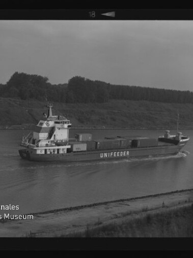 Schwarzweiß-Aufnahme eines Containerschiffs mit der Aufschrift UNIFEEDER, das auf einem ruhigen Fluss oder Kanal fährt; auf dem Deck mehrere Container, im Hintergrund Ufer mit Gras und Baumreihe sowie sichtbarer Filmrand und Museumslogo unten links.