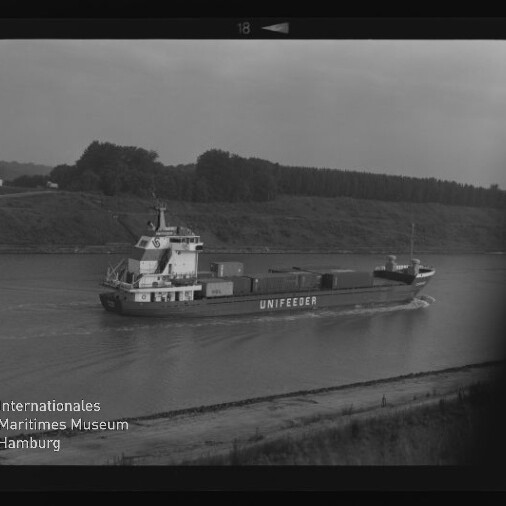 Schwarzweiß-Aufnahme eines Containerschiffs mit der Aufschrift UNIFEEDER, das auf einem ruhigen Fluss oder Kanal fährt; auf dem Deck mehrere Container, im Hintergrund Ufer mit Gras und Baumreihe sowie sichtbarer Filmrand und Museumslogo unten links.
