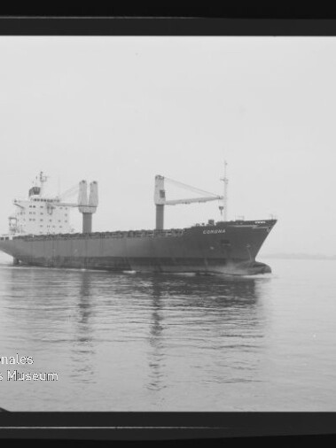 Schwarz-weiß-Foto eines Frachtschiffs mit zwei sichtbaren Ladekränen, das ruhig auf spiegelndem Wasser fährt und dessen Spiegelung zu sehen ist; unten links das Wasserzeichen des Internationalen Maritimen Museums Hamburg.