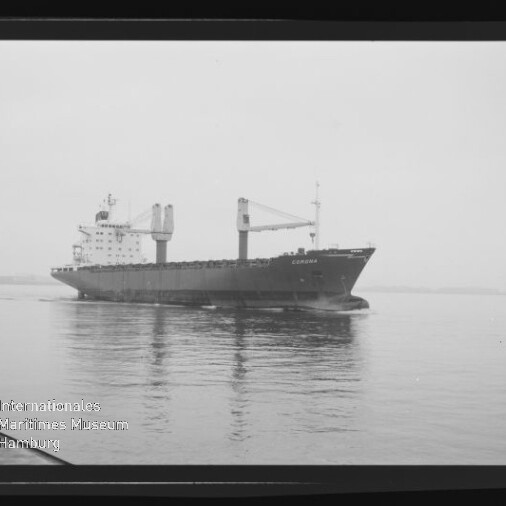 Schwarz-weiß-Foto eines Frachtschiffs mit zwei sichtbaren Ladekränen, das ruhig auf spiegelndem Wasser fährt und dessen Spiegelung zu sehen ist; unten links das Wasserzeichen des Internationalen Maritimen Museums Hamburg.