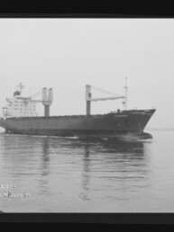 Schwarzweiß-Foto eines großen Frachtschiffs mit zwei markanten Ladekränen und Aufbau am Heck, das ruhig über spiegelndes Wasser fährt, mit sichtbarem Museum-Logo im linken unteren Eck.