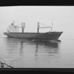 Schwarzweiß-Foto eines Frachtschiffs mit zwei Ladekränen und dem Namen CORONA am Bug, das ruhig über spiegelglattes Wasser fährt, unten links das Logo des Internationalen Maritimen Museums Hamburg sichtbar.