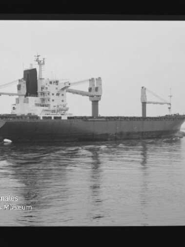 Schwarzweißaufnahme eines Frachtschiffs mit zwei auffälligen Ladekränen und aufragenden Aufbauten in der Bildmitte, das ruhig über spiegelndes Wasser fährt; unten links das Logo und der Schriftzug Internationales Maritimes Museum Hamburg.