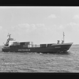 Schwarzweiß-Seitenansicht eines Containerschiffs mit der Beschriftung UNIFEEDER und dem Namen Coronel auf dem Bug, beladen mit Containern und fahrend auf ruhiger See unter leicht bewölktem Himmel; Museumslogo unten links.