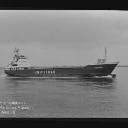Schwarzweiß-Seitenansicht eines Frachtseeschiffs mit großem Schriftzug UNIFEEDER auf dem Rumpf und dem Namen CORONEL am Bug, das bei ruhiger See fährt; unten links sichtbar das Wasserzeichen des Internationalen Maritimen Museums Hamburg.