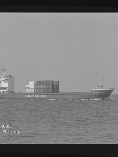 Schwarzweiß-Foto eines UNIFEEDER-Containerschiffs mit mehreren aufgestapelten Containern, das über ruhige See fährt; unten links Logo und Schriftzug des Internationalen Maritimen Museums Hamburg.