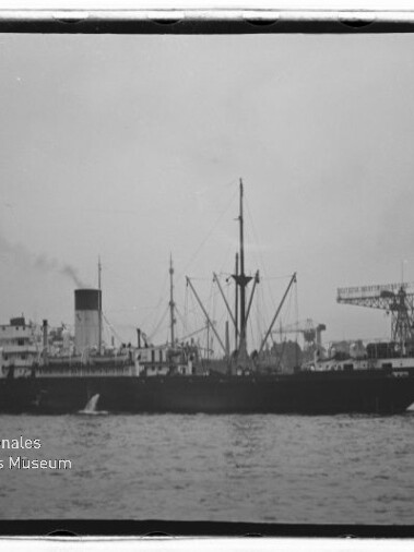 Schwarz-weiß-Foto eines dampfbetriebenen Frachters in Seitenansicht im Hafen mit hohem Schornstein und mehreren Masten, Hafenkränen im Hintergrund und Wasser im Vordergrund; Museumslogo unten links.