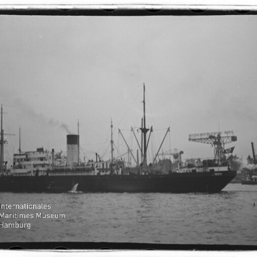 Schwarz-weiß-Foto eines dampfbetriebenen Frachters in Seitenansicht im Hafen mit hohem Schornstein und mehreren Masten, Hafenkränen im Hintergrund und Wasser im Vordergrund; Museumslogo unten links.