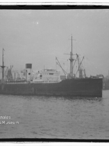 Großes schwarz-weißes Frachtschiff mit Schornstein und Ladekränen an Deck liegt im Hafen auf ruhiger See; im Hintergrund sind weitere Hafenkräne und Industrieanlagen zu sehen, links ein kleines Schlepperboot und unten links ein Wasserzeichen des Internationalen Maritimen Museums Hamburg.