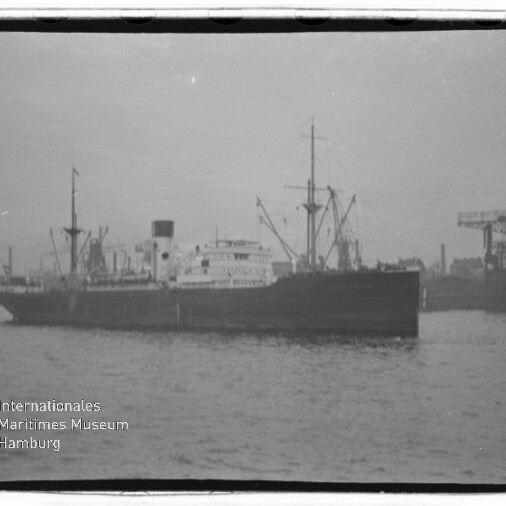 Großes schwarz-weißes Frachtschiff mit Schornstein und Ladekränen an Deck liegt im Hafen auf ruhiger See; im Hintergrund sind weitere Hafenkräne und Industrieanlagen zu sehen, links ein kleines Schlepperboot und unten links ein Wasserzeichen des Internationalen Maritimen Museums Hamburg.