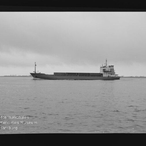 Schwarzweißaufnahme eines langen Frachtschiffs in Seitenansicht auf ruhiger See vor einem tiefen, bewölkten Himmel; unten links ist das Logo des Internationalen Maritimen Museums Hamburg zu sehen.