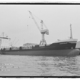 Schwarzweißaufnahme des Frachtschiffs CORSAR, das am Kai liegt, unter einem hohen Hafenkran vor Lagerhallen, mit ruhiger Wasserfläche im Vordergrund und einem Wasserzeichen des Internationalen Maritimen Museums Hamburg in der Ecke.