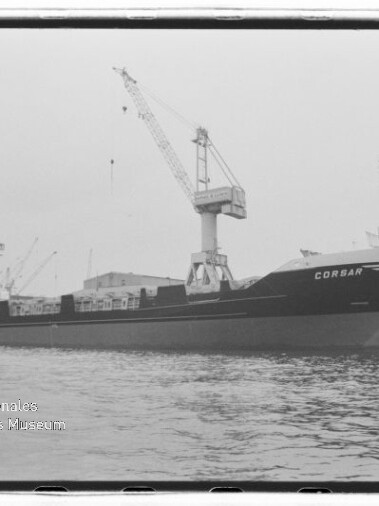 Schwarzweißaufnahme des Frachtschiffs CORSAR, das am Kai liegt, unter einem hohen Hafenkran vor Lagerhallen, mit ruhiger Wasserfläche im Vordergrund und einem Wasserzeichen des Internationalen Maritimen Museums Hamburg in der Ecke.