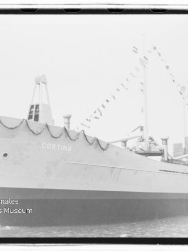 Schwarz-weiß-Foto eines angedockten Frachtschiffs mit dem Namen CORTINA, geschmücktem Bug mit Girlanden und Wimpelketten an Mast und Aufbau, längsseits am Kai vor Hafenbauten.