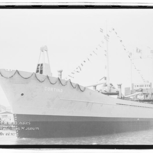 Schwarz-weiß-Foto eines angedockten Frachtschiffs mit dem Namen CORTINA, geschmücktem Bug mit Girlanden und Wimpelketten an Mast und Aufbau, längsseits am Kai vor Hafenbauten.