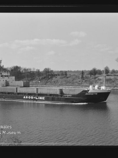 Schwarzweißaufnahme eines Containerschiffs namens CORVETTE mit der Aufschrift AROS-LINE am Rumpf, das mit Containern beladen einen Kanal entlangfährt; im Hintergrund Ufer mit Bäumen und unten links ein Museumswasserzeichen.
