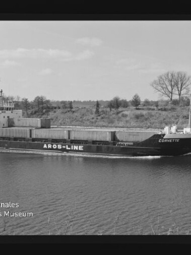 Schwarzweiß-Foto eines Containerschiffs namens „Corvette“ mit großer Aufschrift „AROS-LINE“, das ruhig einen Kanal passiert; im Hintergrund flaches Ufer mit Bäumen, links unten Museumssignet und sichtbarer Filmrahmenrand.