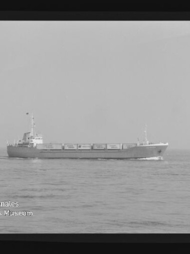 Schwarzweißaufnahme eines mittleren Frachtschiffs, das auf ruhiger See vor einem flachen Horizont entlangfährt; am linken unteren Bildrand ist das Logo des Internationalen Maritimen Museums Hamburg sichtbar.