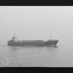 Schwarzweißaufnahme eines Frachtschiffs mit Containern, das bei ruhiger, nebliger See von links nach rechts fährt; Filmrand und Wasserzeichen des Internationalen Maritimen Museums Hamburg sichtbar.